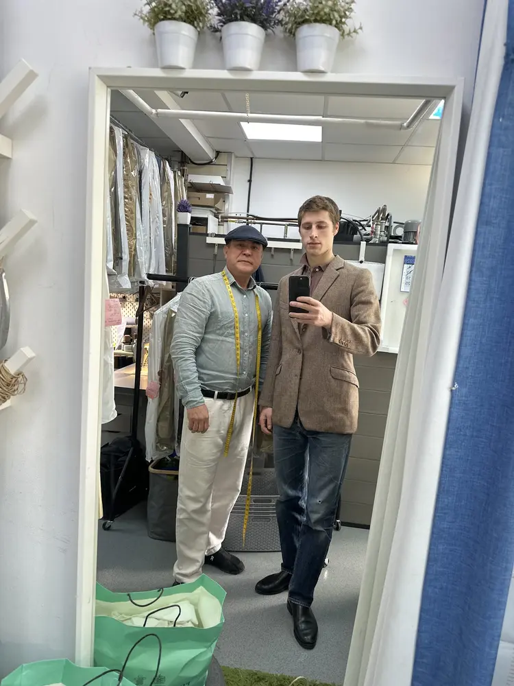 Mr. Ahmad, tape measure round his neck, with a customer in a tweed jacket at the fitting mirror